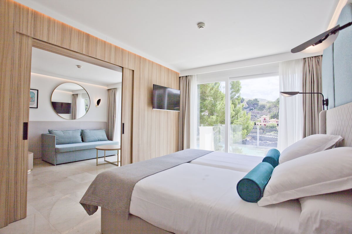 Habitación de hotel moderna con cama, sofá, televisor y un amplio ventanal con vistas al exterior.