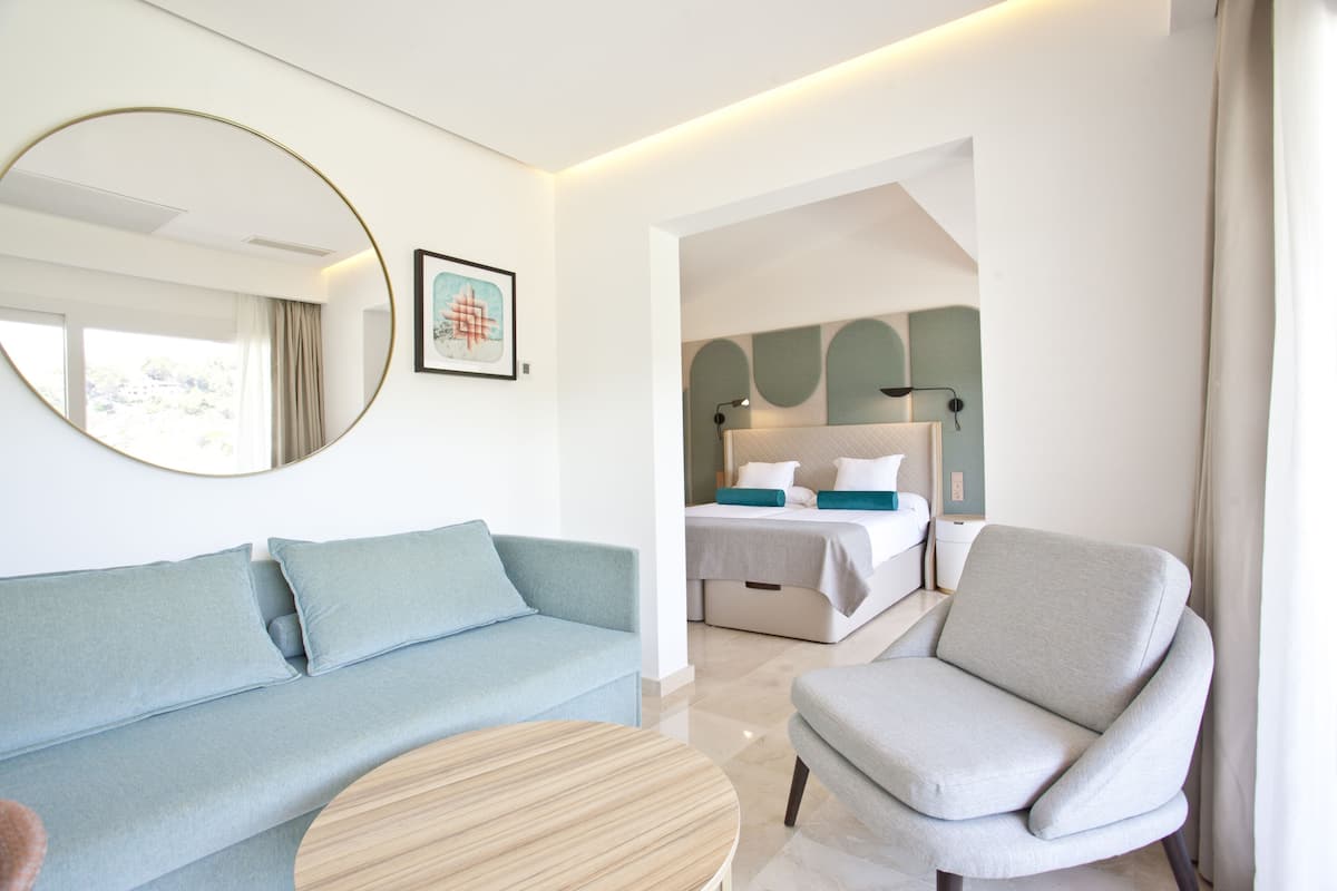 Habitación de hotel moderna con un sofá azul claro, una mesa redonda de madera y un sillón gris. Un espejo grande y un cuadro decoran la pared.