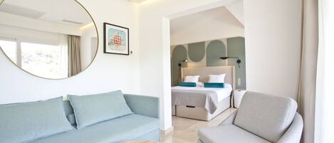 Ett modernt hotellrum med en ljusblå soffa, ett runt träbord och en grå fåtölj. En stor spegel och en inramad tavla pryder väggen.