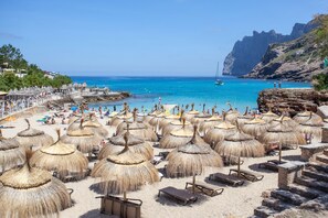 Una playa con numerosas sombrillas de paja y tumbonas, un mar azul cristalino y acantilados rocosos al fondo.