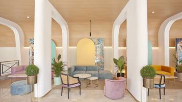 A spacious lobby with a modern design, featuring a central seating area with sofas and chairs, surrounded by potted plants and a large abstract painting on the wall.