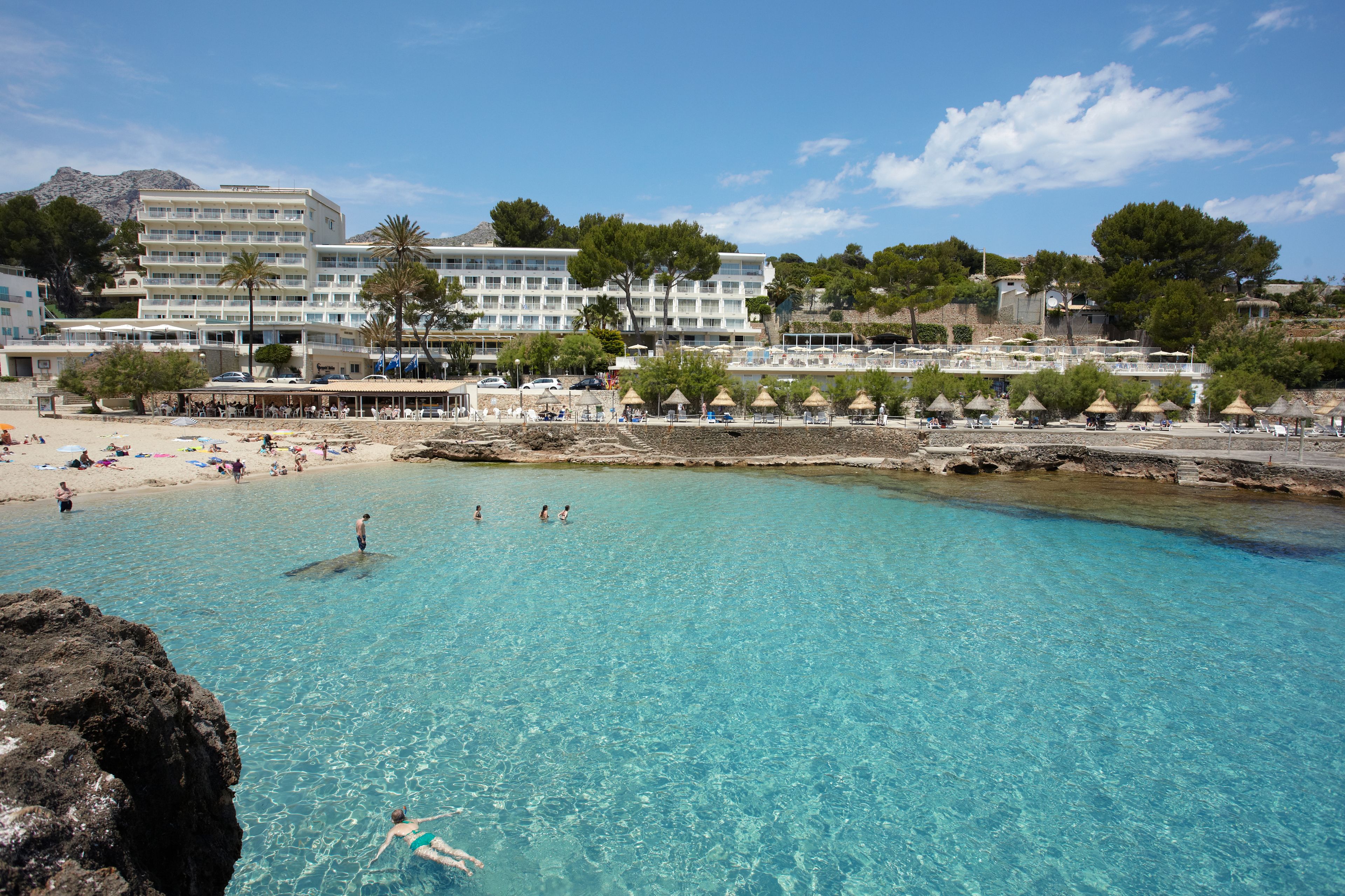 Un hotel costero con varios pisos, una zona de playa con sombrillas y agua turquesa cristalina.