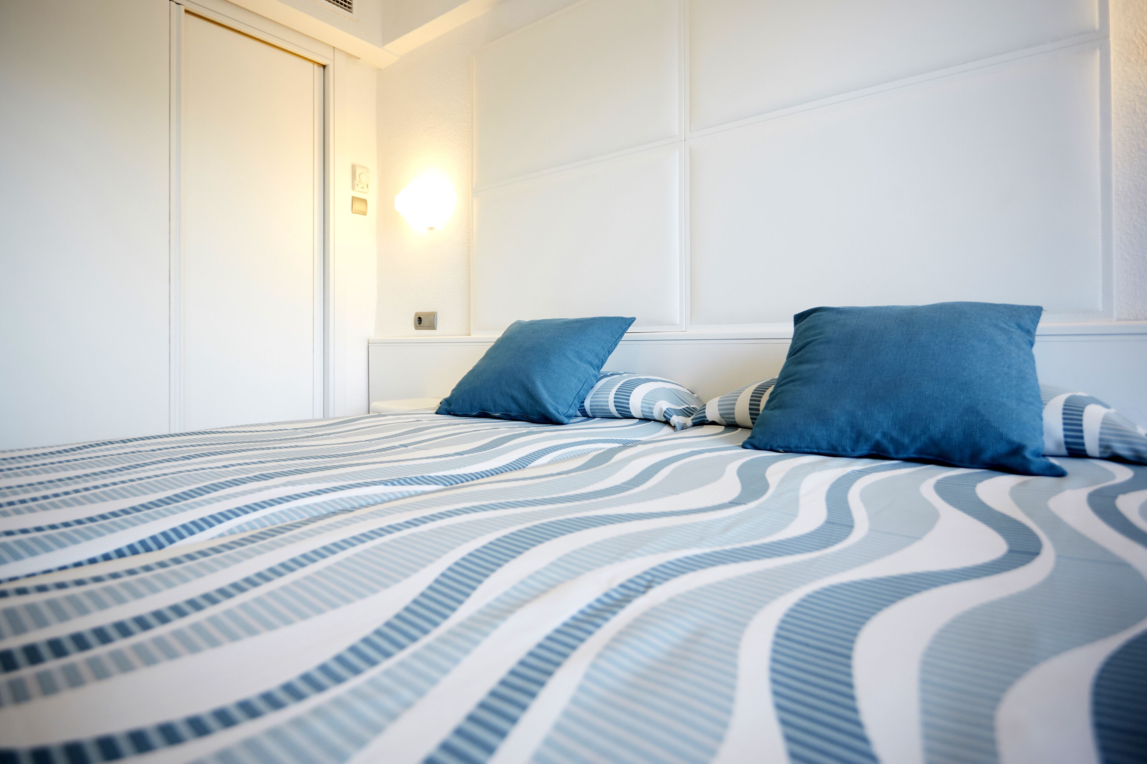 A hotel room with a bed featuring a blue and white striped bedspread, two blue pillows, and a wall-mounted light fixture.