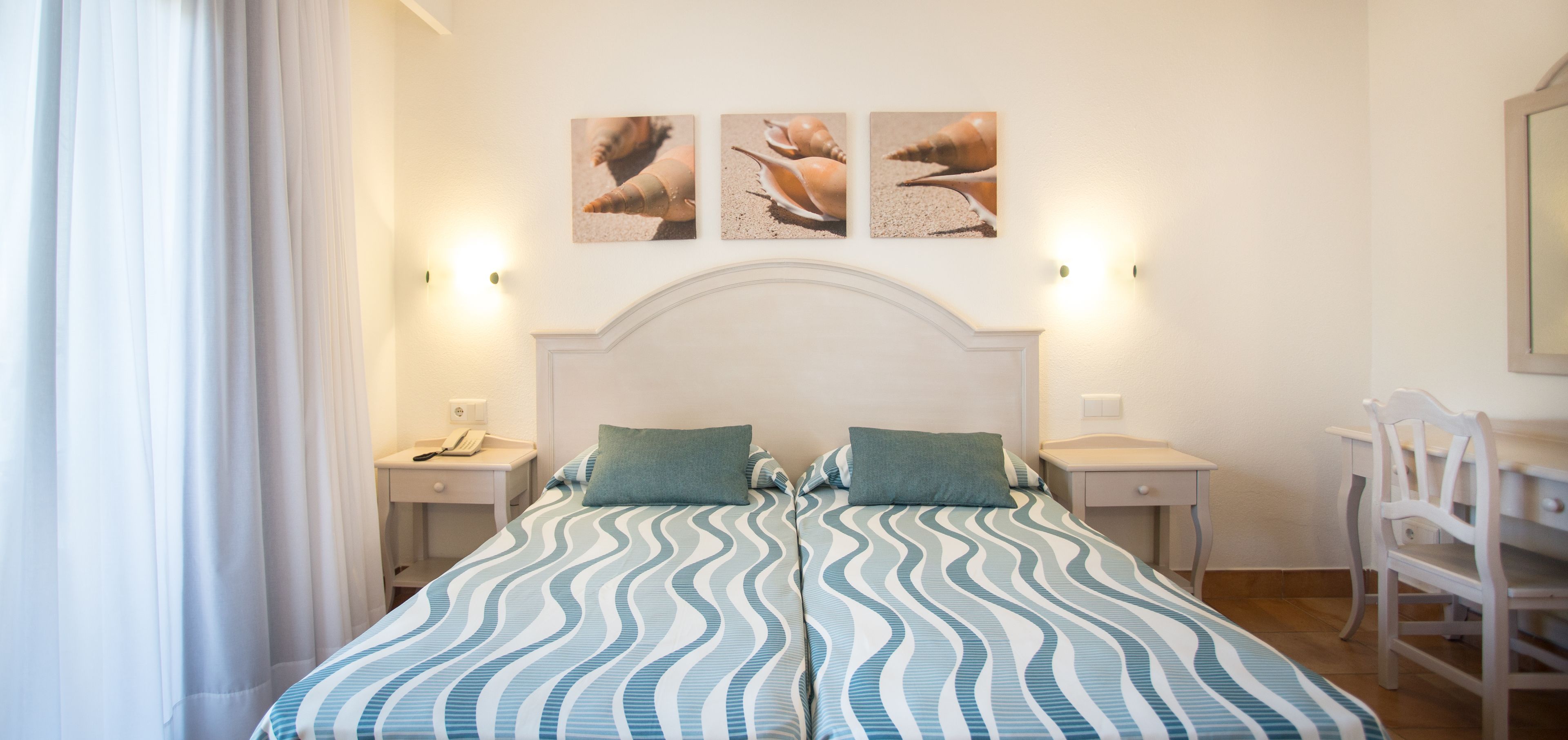 A bedroom with a bed, two bedside tables, a desk, and a chair. There are three wall-mounted pictures above the bed.