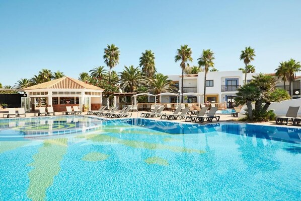 A swimming pool with lounge chairs, palm trees, and a building with a tiled roof. - Insotel Hotel Formentera Playa (Formentera)