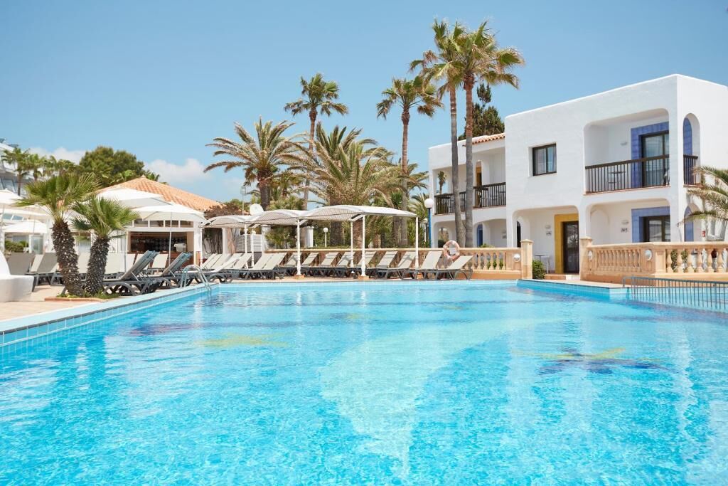 A swimming pool with lounge chairs and umbrellas, surrounded by palm trees and a white building with balconies.