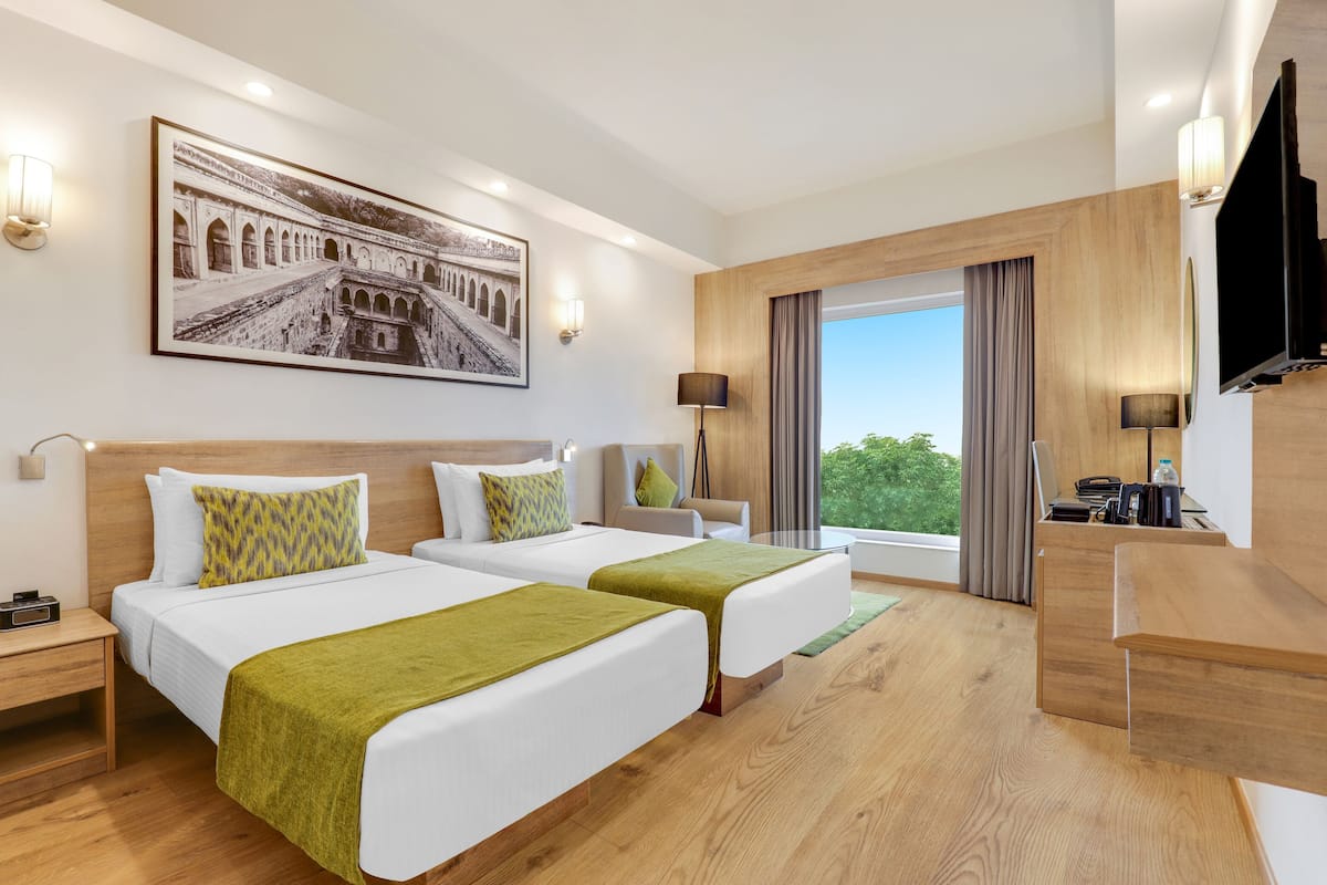 A hotel room with two beds, a desk, a TV, and a large window with a view of greenery.