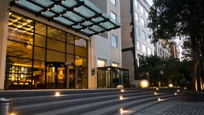 A modern hotel entrance with a glass facade, outdoor seating area, and a staircase leading to the entrance. - Lemon Tree Premier City Center Gurugram (Gurugram)