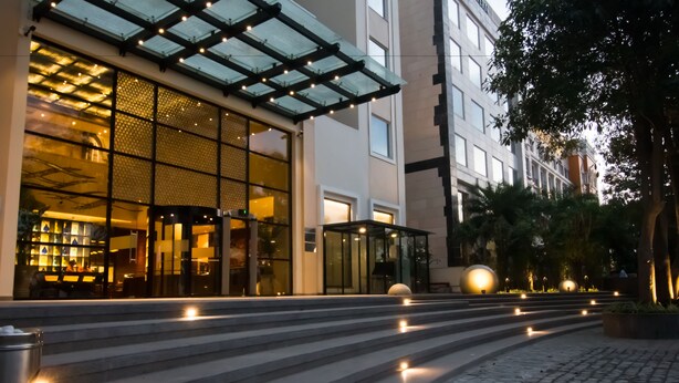 A modern hotel entrance with a glass facade, outdoor seating area, and a staircase leading to the entrance. - Lemon Tree Premier City Center Gurugram (Gurugram)