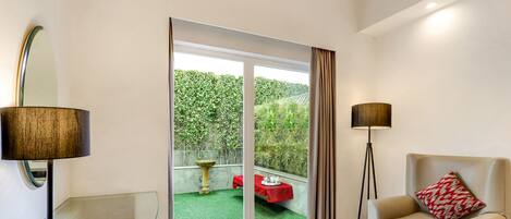 A modern living room with a glass coffee table, a sofa with a patterned pillow, a floor lamp, and a sliding glass door leading to a balcony with a red table and a garden view.