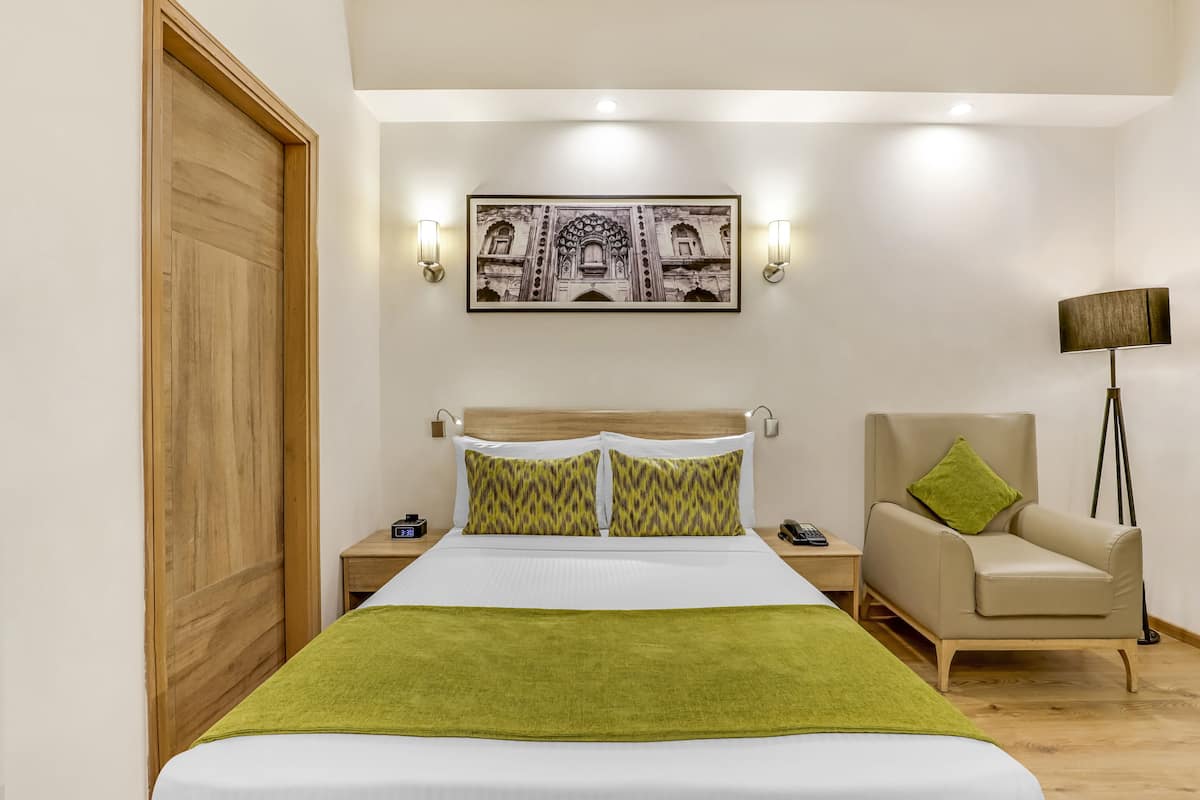 A hotel room with a bed, a chair, a nightstand, a lamp, and a wall-mounted picture.