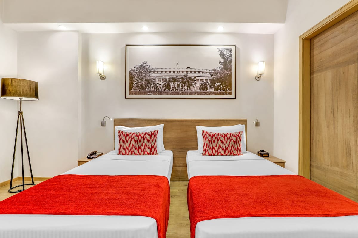 A hotel room with two beds, each with a red blanket, white bedding, and matching red and white pillows. There is a framed picture on the wall above the beds, a floor lamp, and a wooden door.