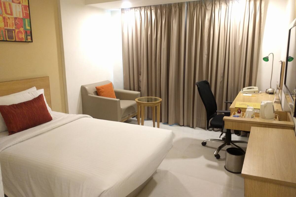 A hotel room with a large bed, a desk with a telephone and lamp, a chair, and a small table.