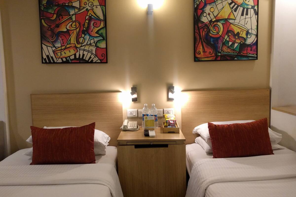 A hotel room with two beds, each with red pillows, a wooden headboard, a small table with a phone, a water bottle, and snacks. There are two wall-mounted light fixtures and two colorful abstract paintings above the beds.