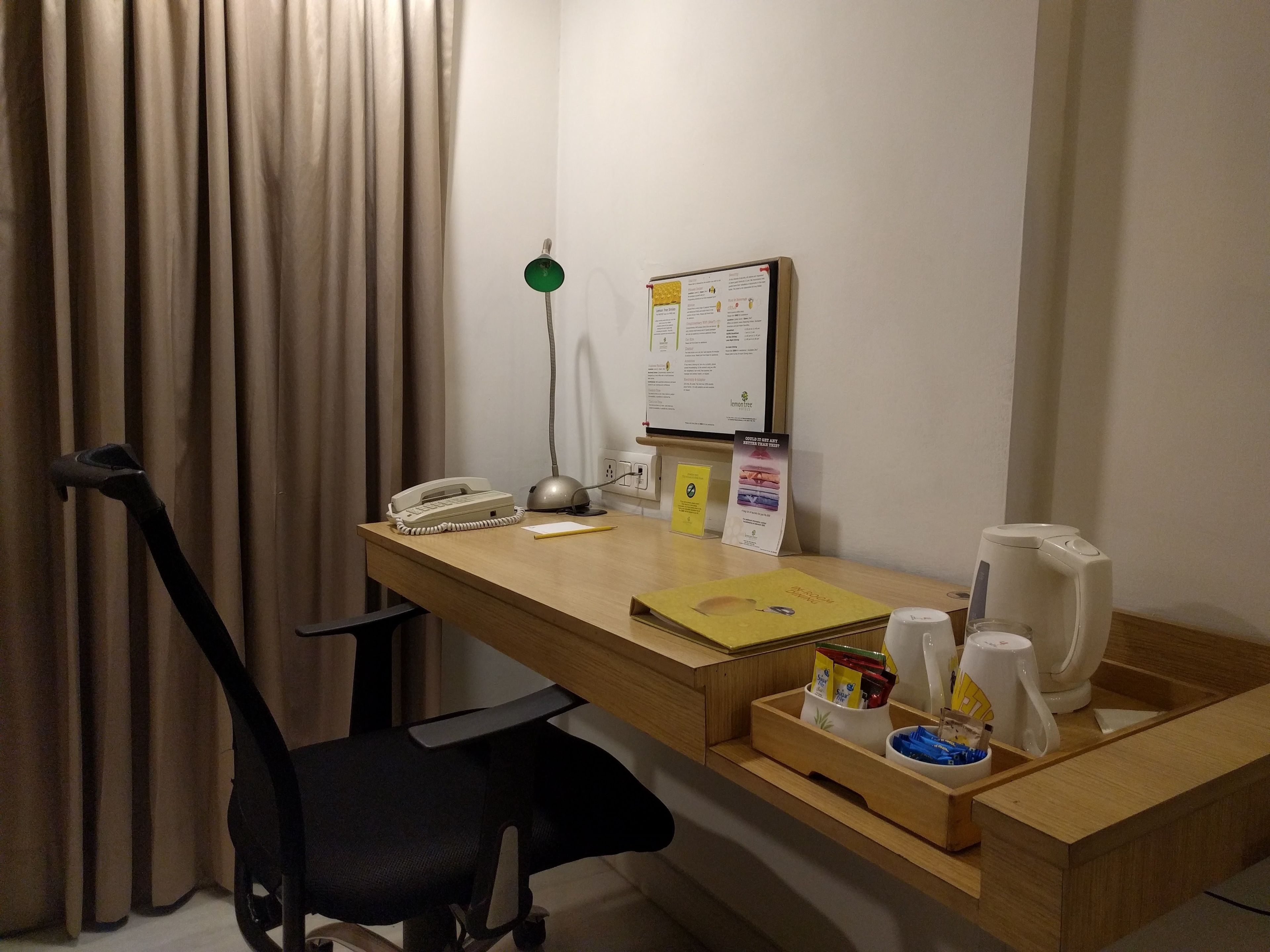 A hotel room with a wooden desk, a telephone, a lamp, a bulletin board, and a coffee maker.