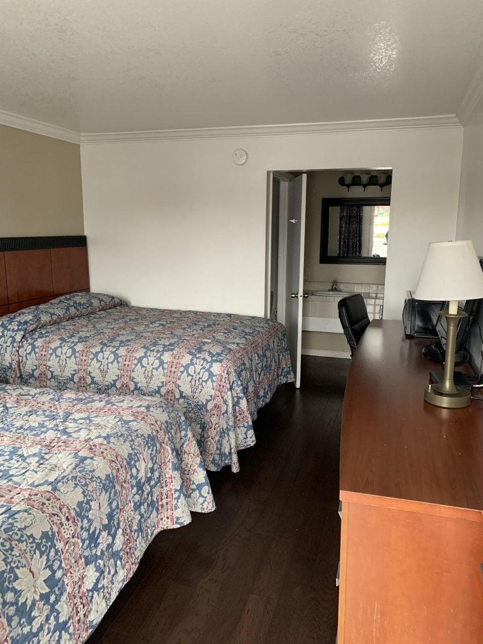 A hotel room with two beds, a desk, a lamp, and a bathroom visible through an open door.