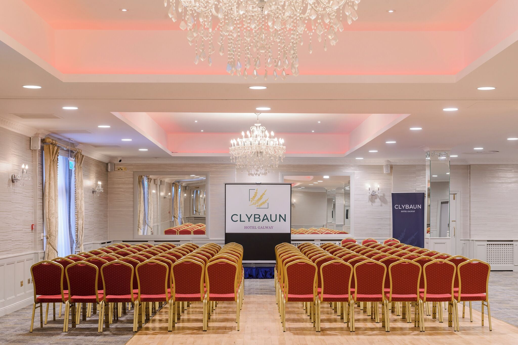 Un salón de conferencias con filas de sillas rojas, una araña de cristal y un cartel del Hotel Clybawn Galway.