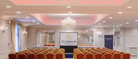 Un salón de conferencias con filas de sillas rojas, una araña de cristal y un cartel del Hotel Clybawn Galway.