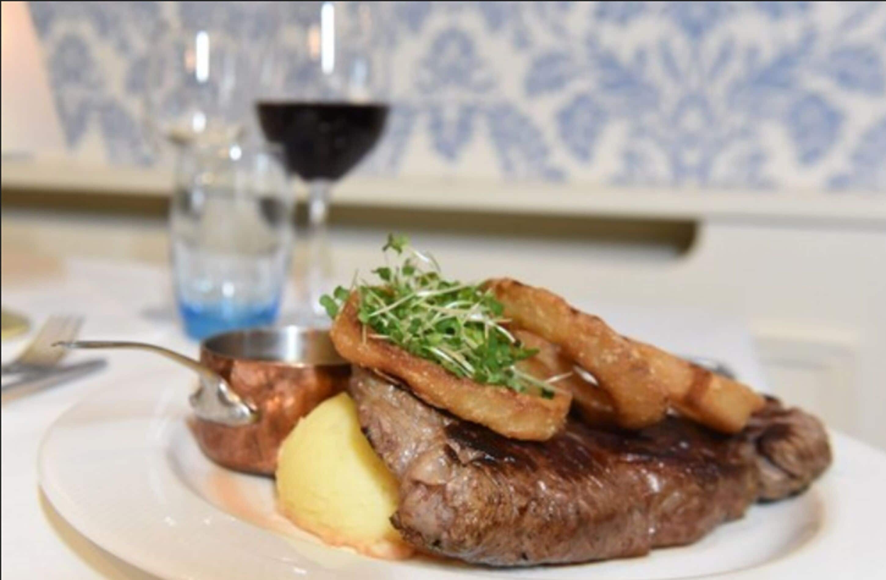 A plate with a steak, fried potatoes, and a small portion of greens, accompanied by a glass of red wine and a copper pot.