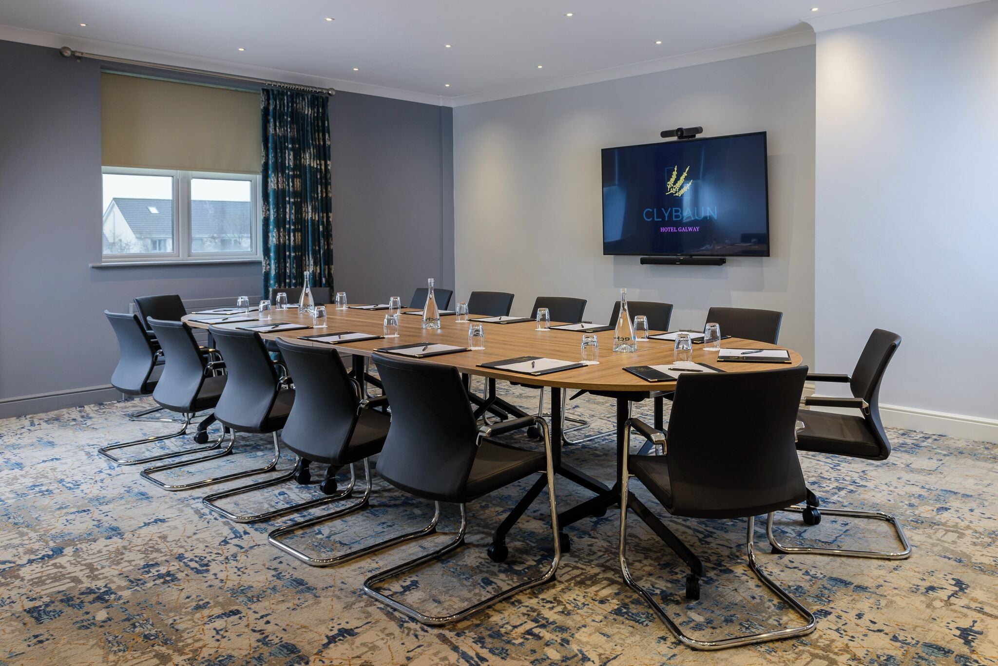Een vergaderzaal met een grote tafel, stoelen, een televisiescherm en een raam met gordijnen.