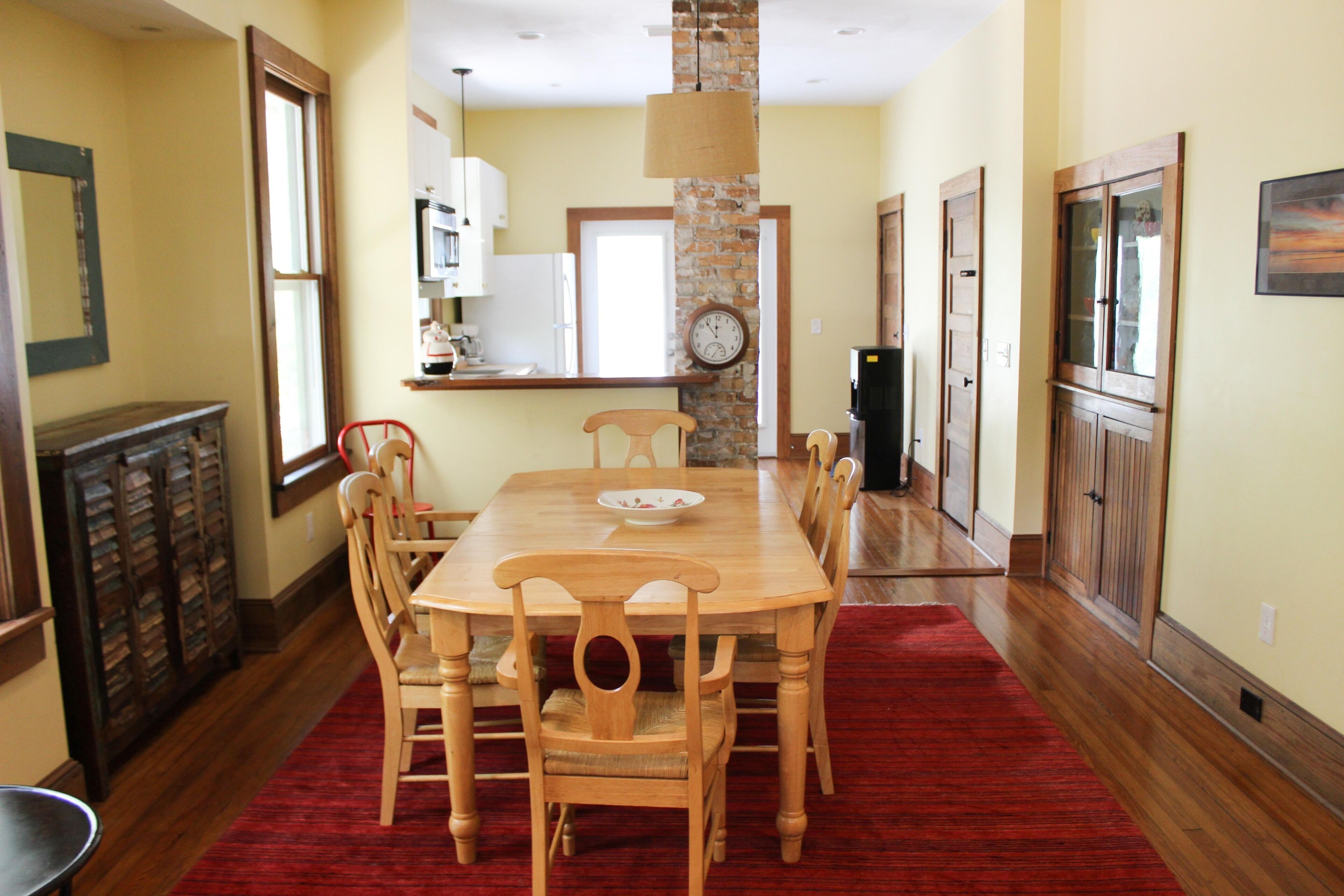 A dining area with a wooden table, chairs, a red rug, a clock on the wall, and kitchen cabinets in the background.