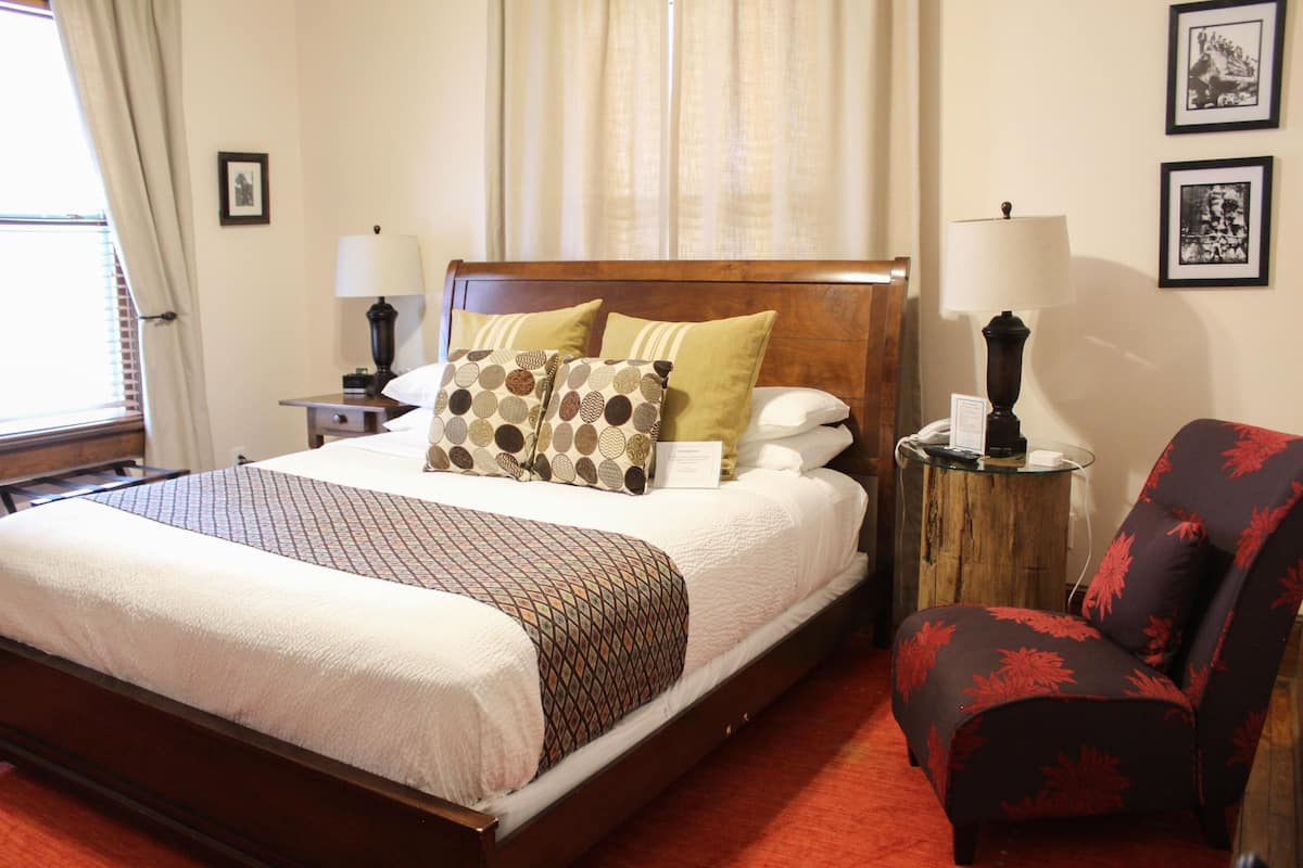 A neatly made bed with patterned pillows, a wooden headboard, a side table with a lamp, a chair with a floral pattern, and two framed pictures on the wall.