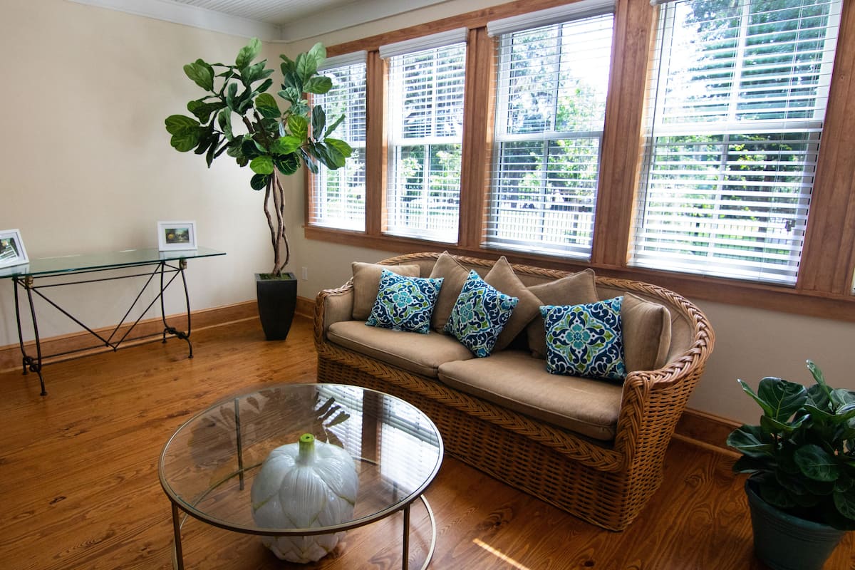 A living room with a wicker sofa, a glass coffee table, a potted plant, and a view of the outdoors through large windows.