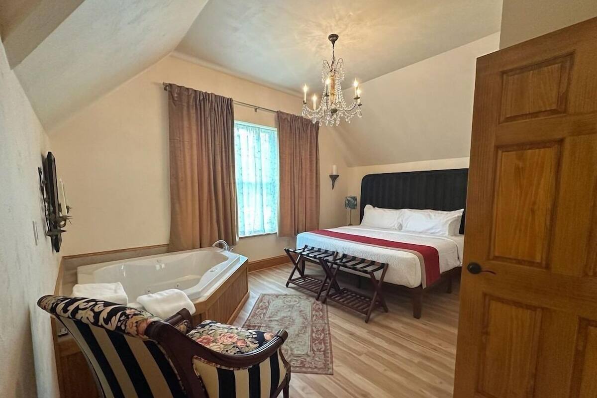 A bedroom with a bed, a wooden door, a chandelier, a bathtub, and a chair.