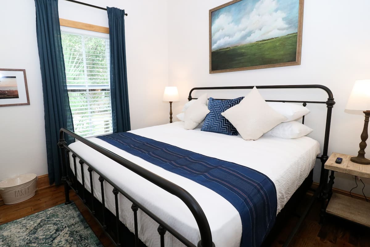 A neatly made bed with a blue and white bedspread, a metal headboard, two bedside lamps, a framed picture on the wall, and a window with curtains.