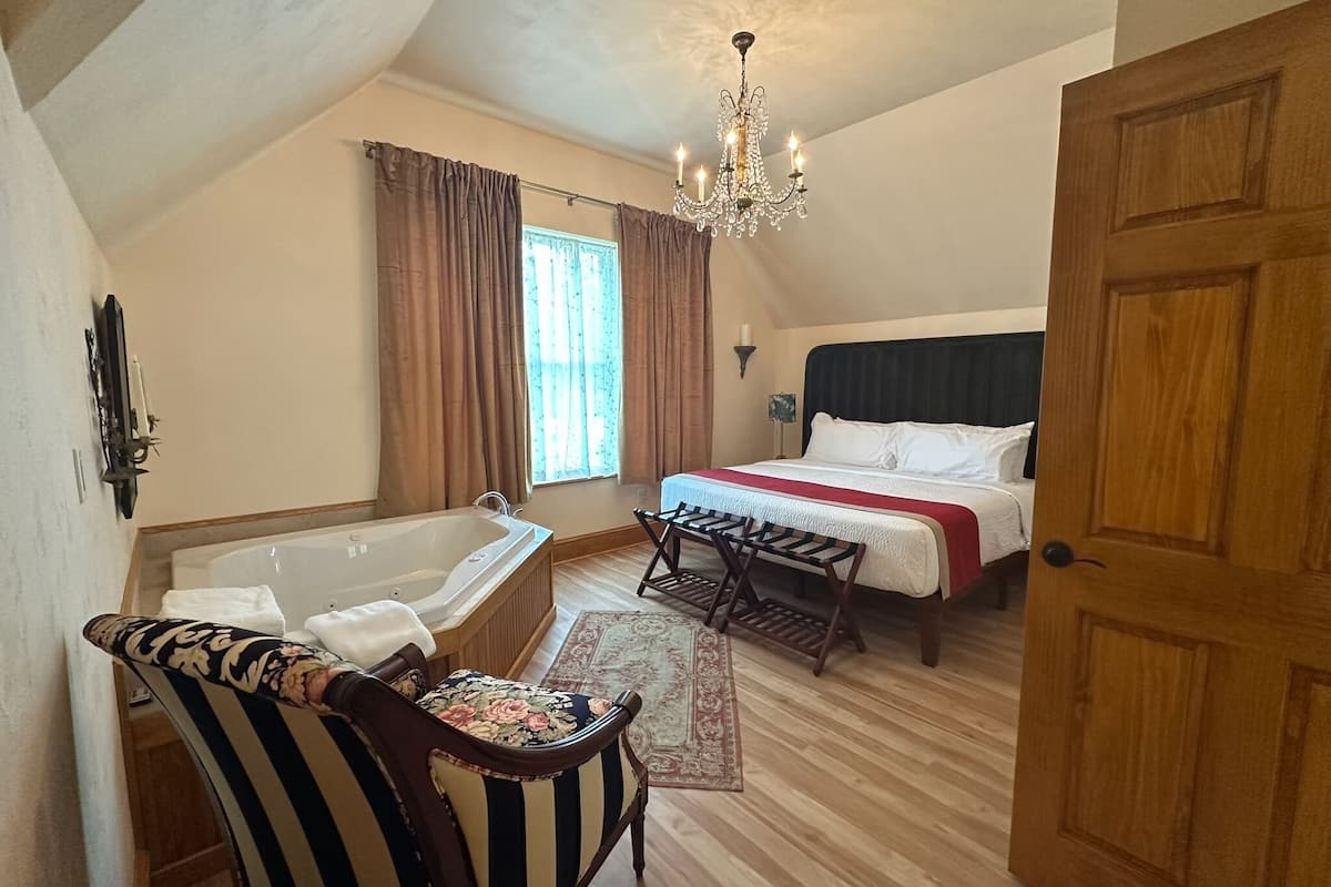 A bedroom with a bed, a wooden door, a chandelier, a window with curtains, a bathtub, and a chair.
