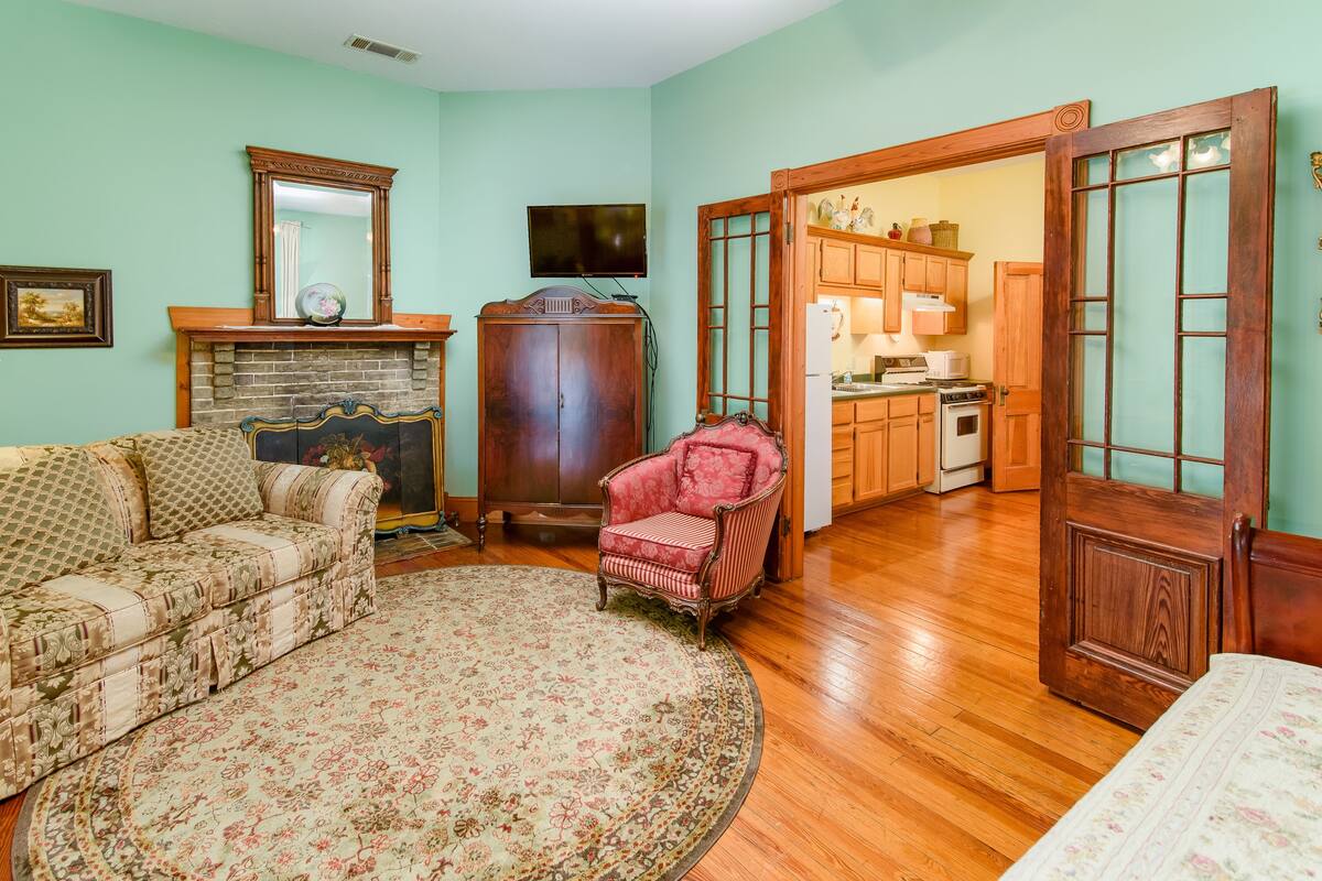 A living room with a fireplace, a sofa, a chair, and a kitchen visible through an open door.