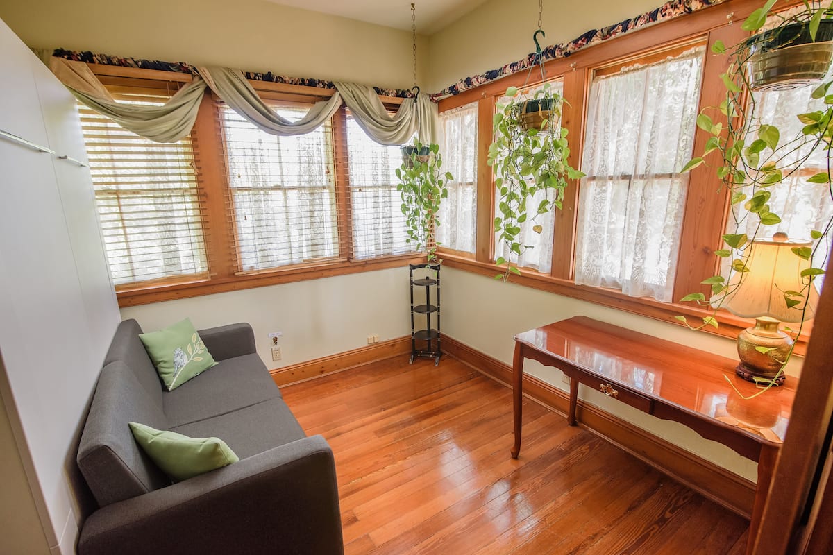 A room with a wooden floor, a grey sofa with green cushions, a wooden table, and windows with curtains and hanging plants.
