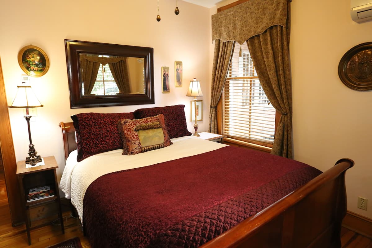 A bedroom with a large bed, a nightstand, a lamp, a mirror, a window with curtains, and wall decorations.
