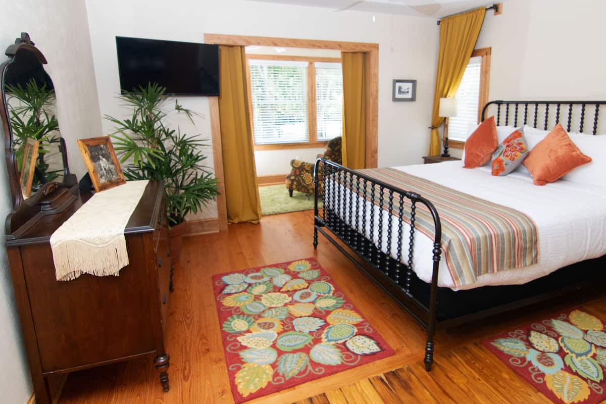 A bedroom with a bed, wooden dresser, TV, potted plants, and a window with curtains.