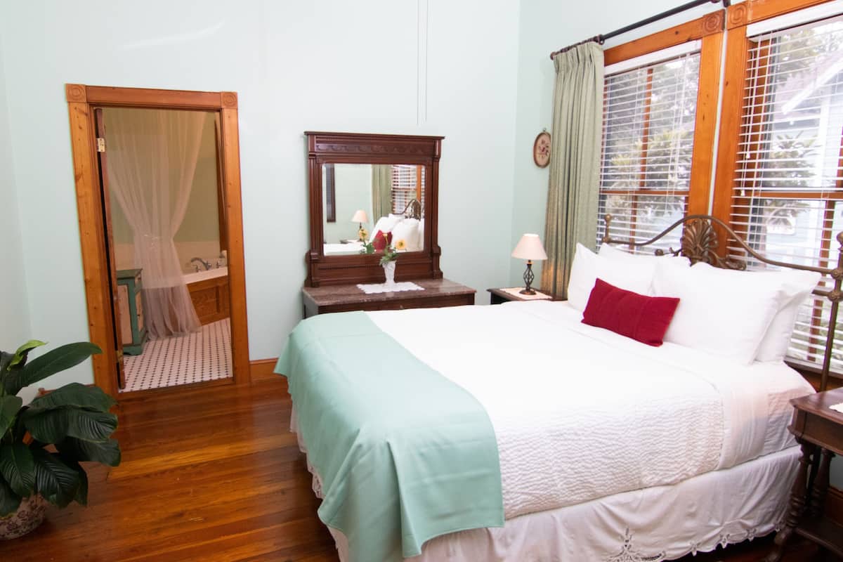 A bedroom with a bed, a nightstand, a mirror, a window with curtains, and a potted plant.