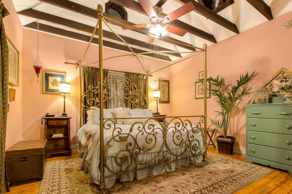 A four-poster bed with a decorative headboard, a green dresser, and a ceiling fan in a room with wooden beams and a pink wall.