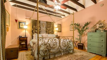 A four-poster bed with a decorative headboard, a green dresser, and a ceiling fan in a room with wooden beams and a pink wall.