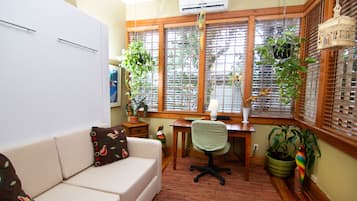 Una habitación acogedora con un sofá blanco, un escritorio de madera y una ventana con persianas. Se observan varias plantas en macetas y una jaula colgante.