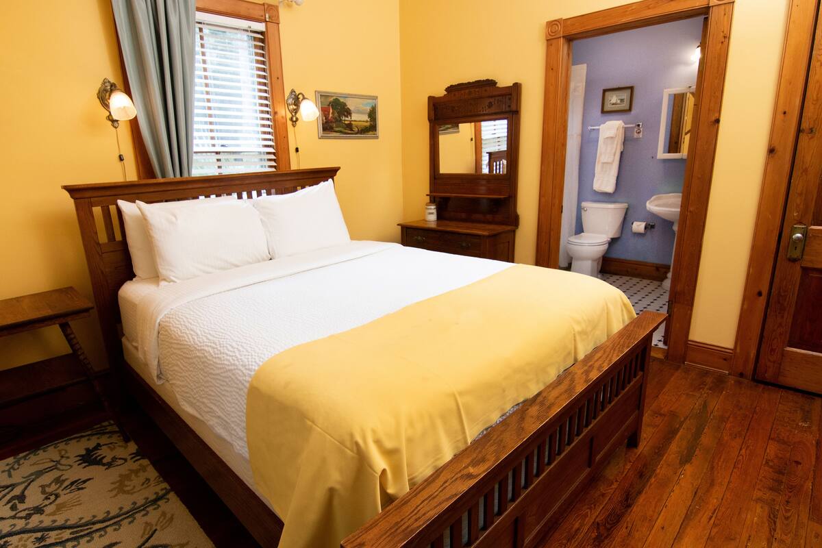 A bedroom with a wooden bed, a nightstand, a bedside table, a window with curtains, a bathroom visible through an open door, and a wooden floor.