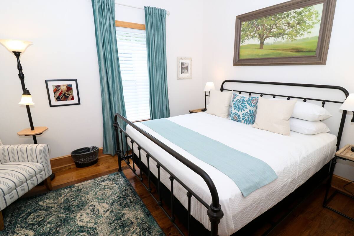 A neatly arranged bedroom with a bed, bedside tables, a lamp, a chair, a rug, and a painting on the wall.
