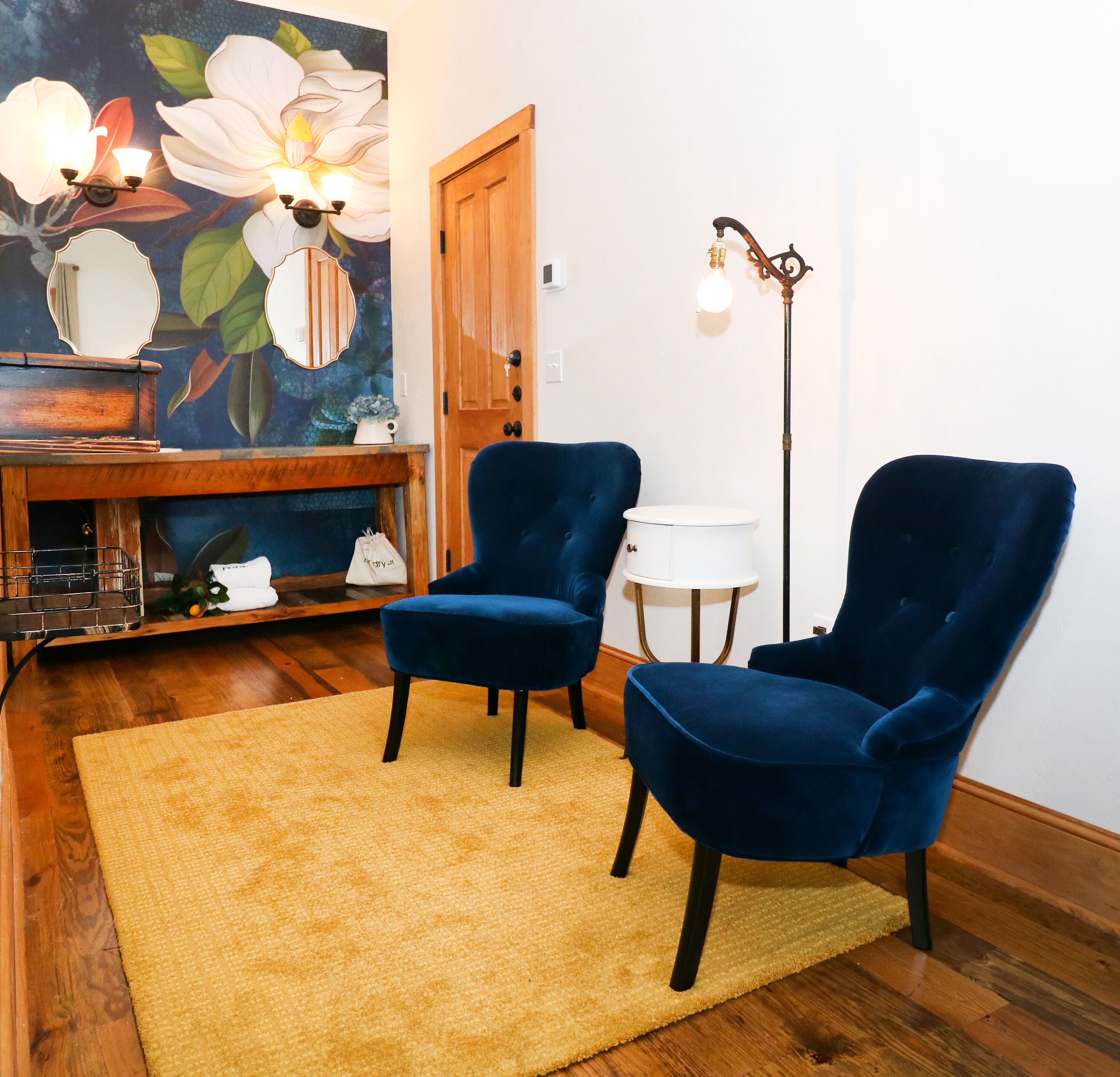 Una habitación con dos sillones azules, suelo de madera, alfombra amarilla, un cuadro de flores, un aparador de madera y una lámpara de pie.