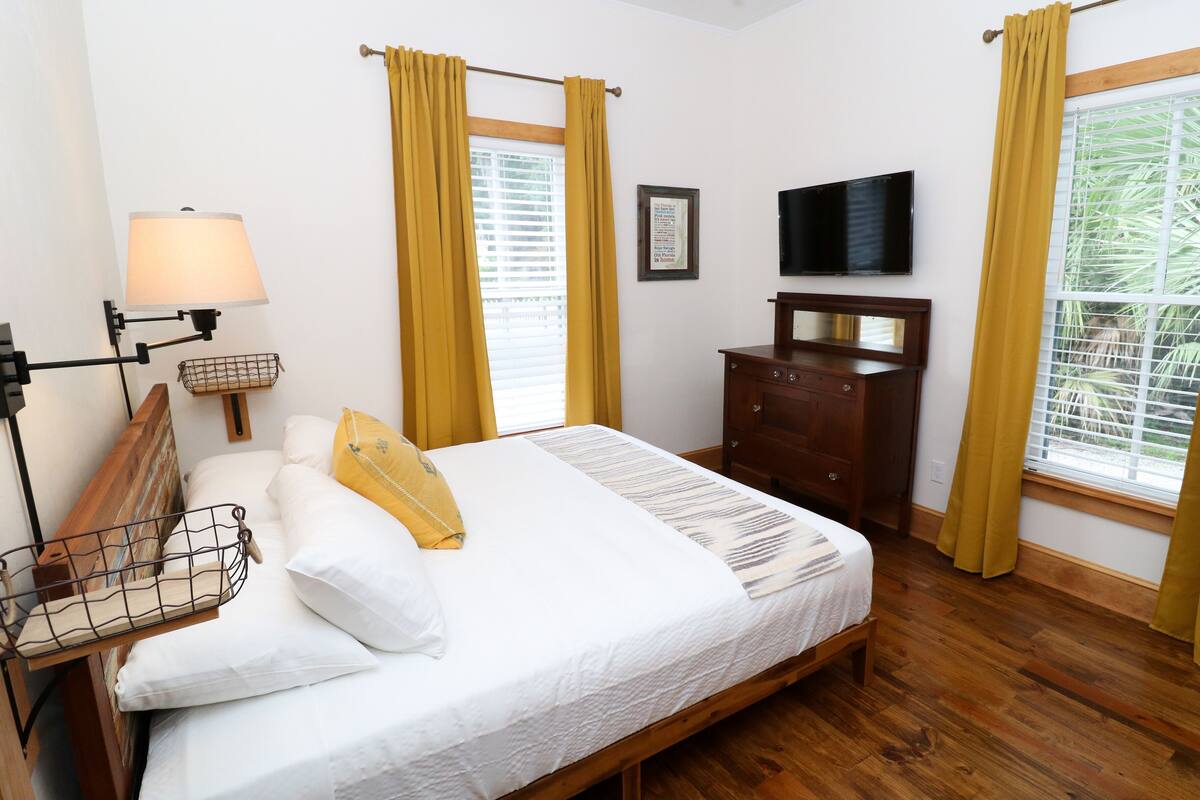 A bedroom with a wooden bed, a nightstand, a lamp, a dresser, a TV, and a window with curtains.