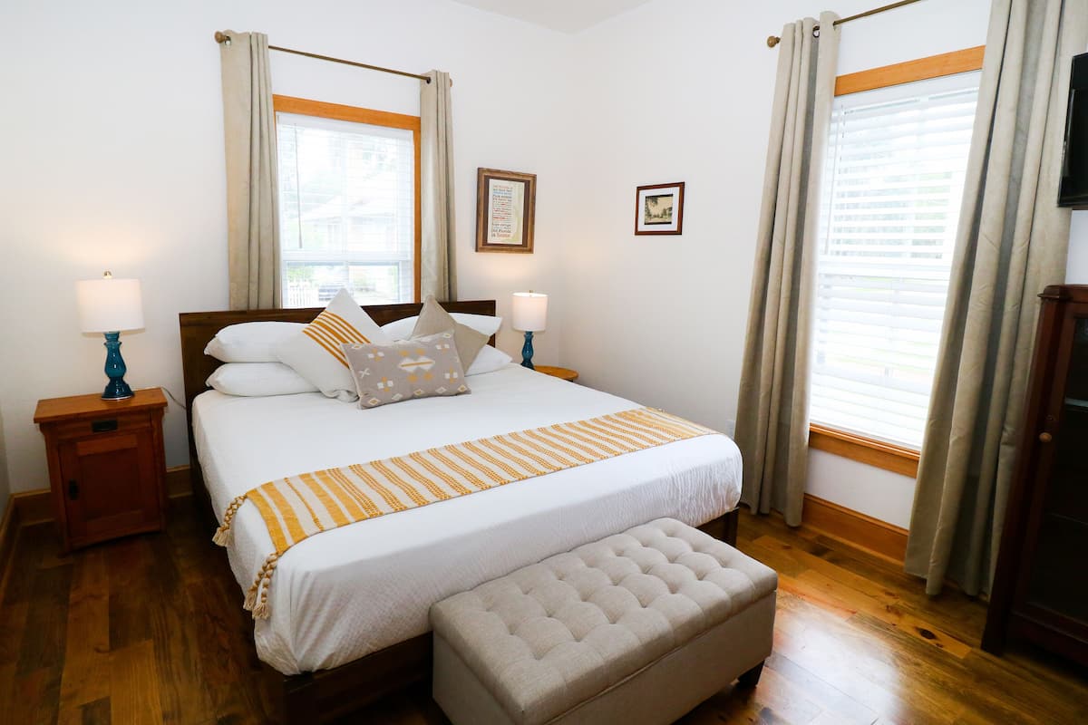 A neatly made bed with a striped blanket, two bedside tables with lamps, a wooden bench, and a television on the wall.
