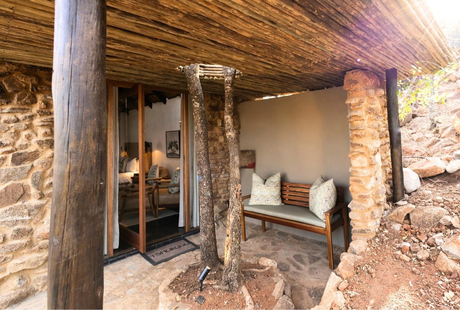 A rustic stone building with a wooden bench and a door leading to a room with a bed and a table.