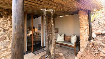 A rustic stone building with a wooden bench and a door leading to a room with a bed and a table.