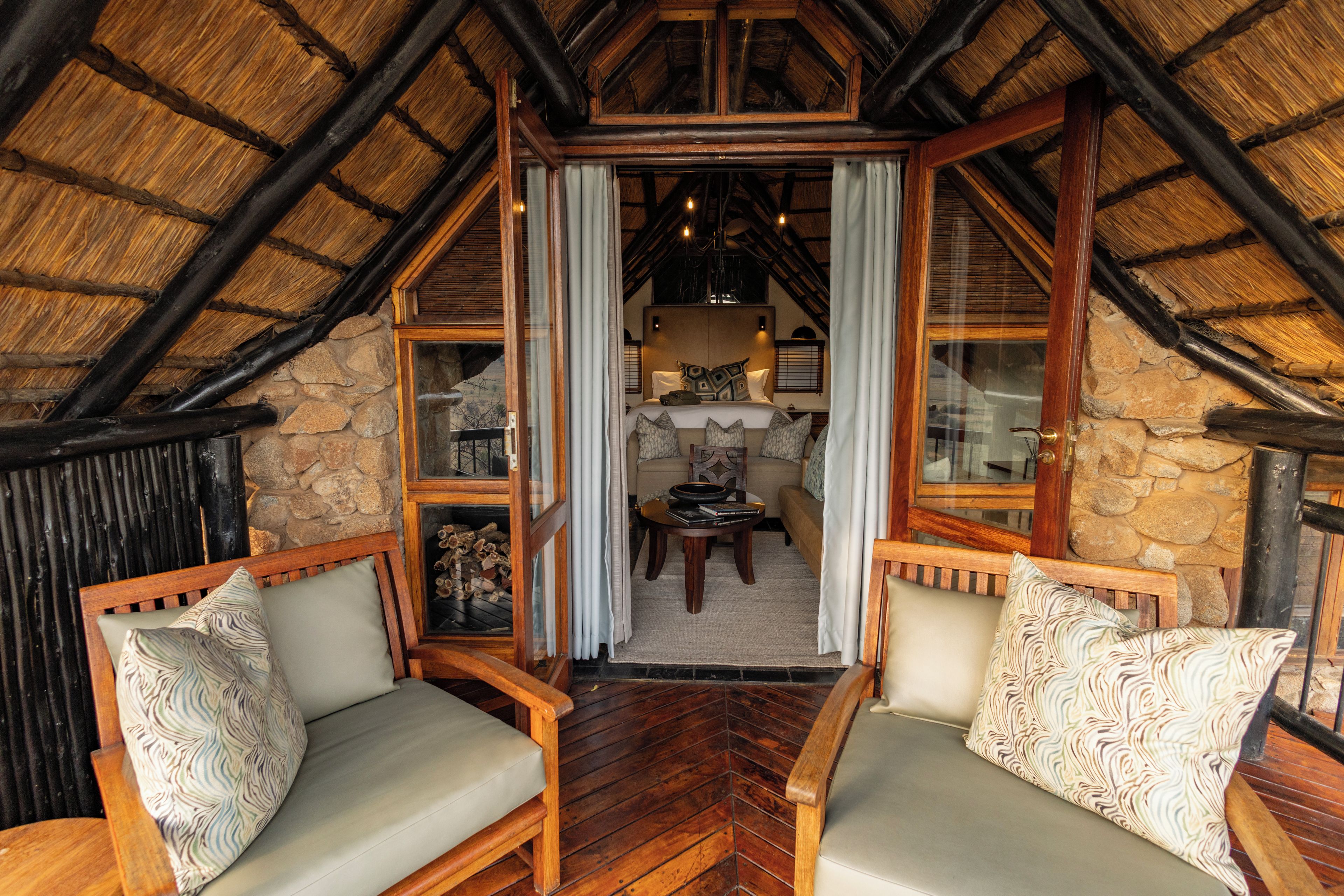 A cozy room with a sloped ceiling, wooden walls, and a stone fireplace. There are two wooden chairs with cushions and a small table with a book and a bowl.