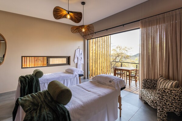 A spa room with two massage tables, a wooden ceiling light, a view of the outdoors, and a patterned sofa.