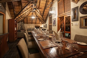 A dining area with a long wooden table set for a meal, a stone wall, and a thatched roof ceiling.