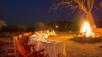 An outdoor dining setup with tables and chairs, a fire pit, and a tree in the background.