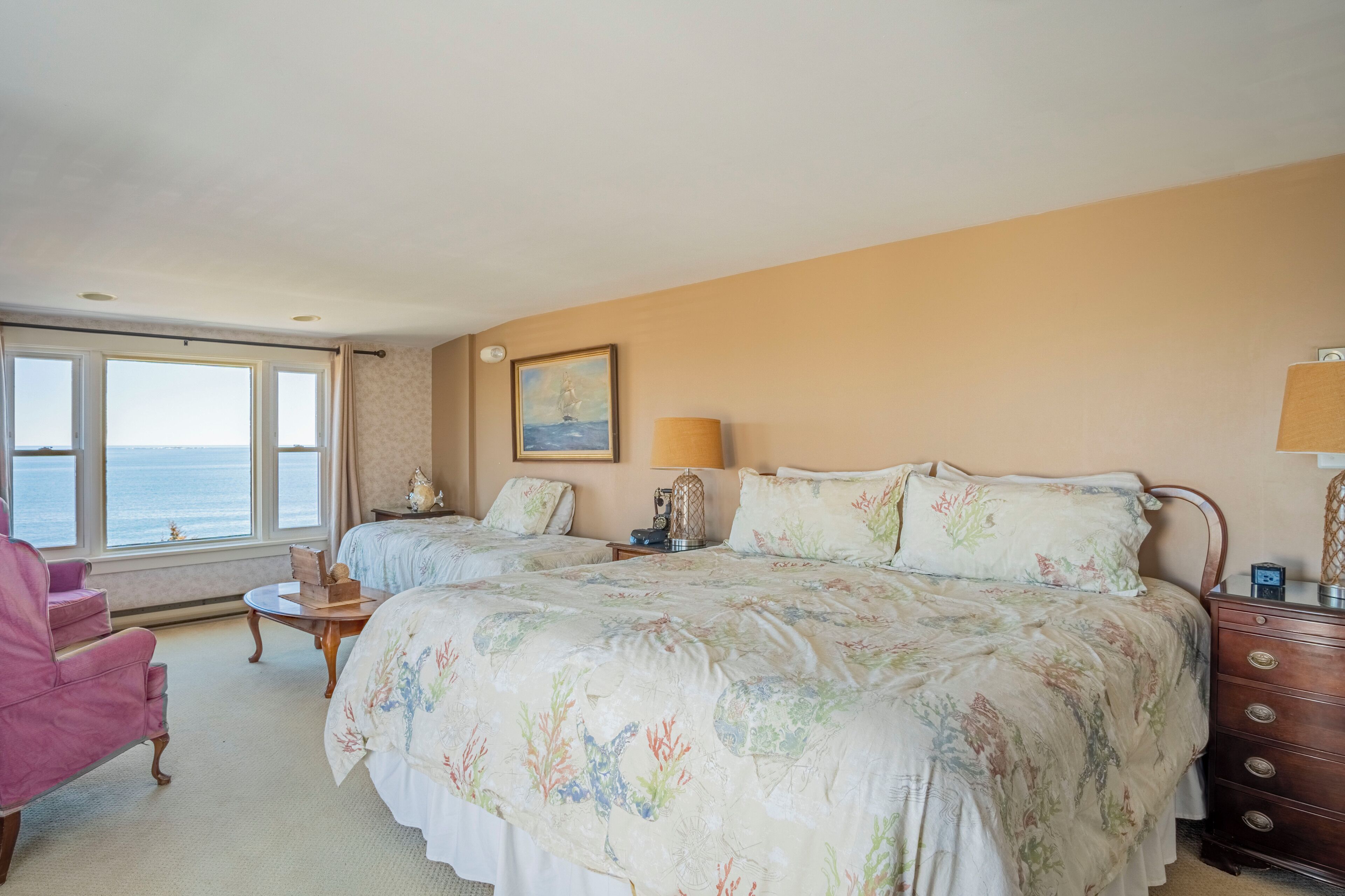 Habitación de hotel con dos camas, vista al mar, una silla rosa y un aparador de madera.